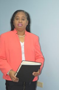 Middle-aged woman of mixed ethnicity, wearing a salmon-coloured jacket, white blouse and black pants. She is holding a binder of documents, and standing in front of a blue wall.