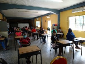 A group of masked people sit physically-distanced at school desks