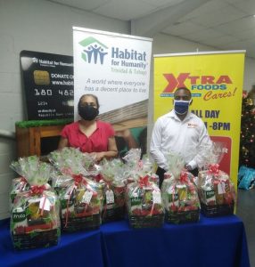 A woman in a red dress and a man in a long-sleeved white shirt stand behind a table  packed with Christmas hampers. Behind them are signs for Habitat for Humanity Trinidad and Tobago and Xtra Foods